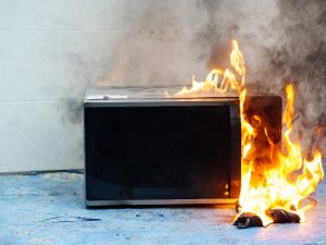 A microwave oven on fire, with flames engulfing the damaged microwave door and a charred object on the floor.