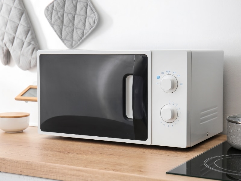 A white, energy efficient refurbished microwave oven sits on a light brown countertop in a kitchen setting.