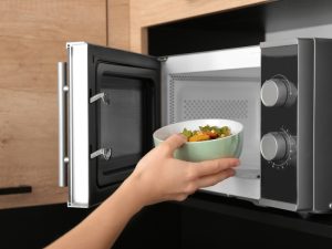 Hand placing a bowl of cooked vegetables into a microwave, demonstrating safe usage and maintenance tips after buying a used microwave for the kitchen.