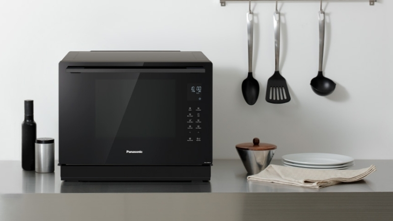 Panasonic NN-CS89LB – Modern black microwave on a sleek countertop, with kitchen utensils, plates, and a decorative bowl nearby